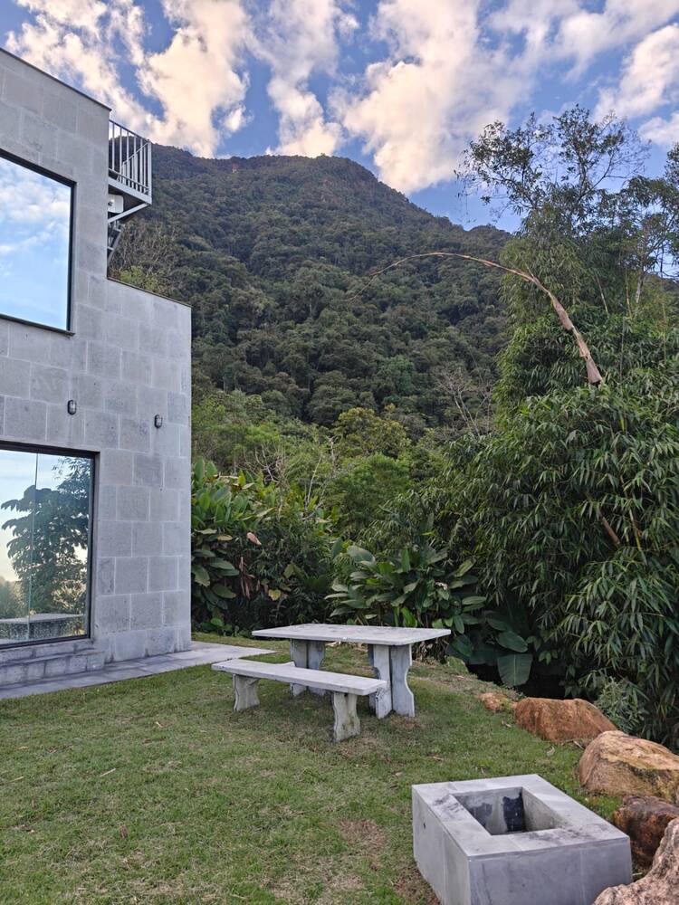 Fachada de blocos de concreto da Casa de Pedra, com as duas janelas de vidro do piso ao teto refletindo o céu e a serra ao fundo.