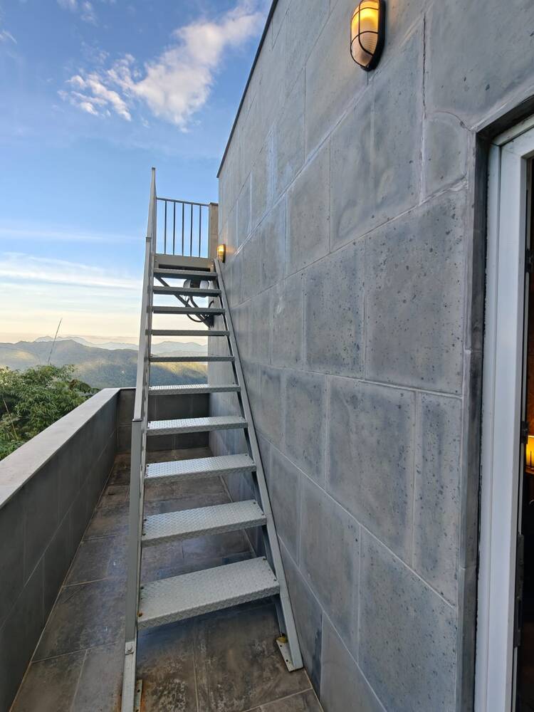 Escada metálica externa que sobe até o terraço no topo da torre, com vista para o vale ao fundo.