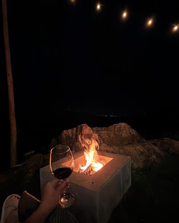 Fogueira acesa em uma base de concreto, com uma taça de vinho tinto em primeiro plano, varal de luzes aceso por cima e as luzes do vale ao longe.