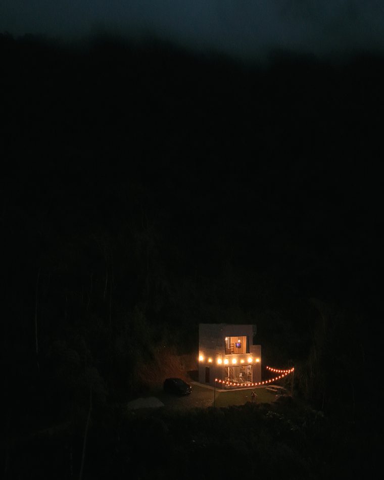 A Casa de Pedra iluminada por dentro à noite, com um varal de luzes contornando o terreno, sozinha no escuro da encosta.