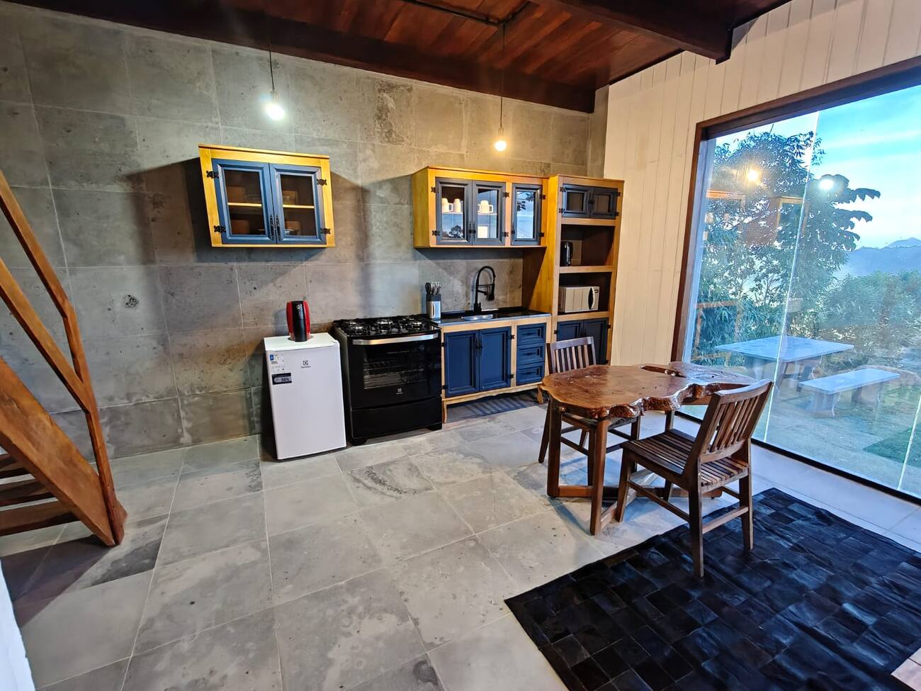 Cozinha compacta com armários azuis, fogão, geladeira pequena e mesa redonda de madeira em frente a uma porta de vidro com vista para a serra.