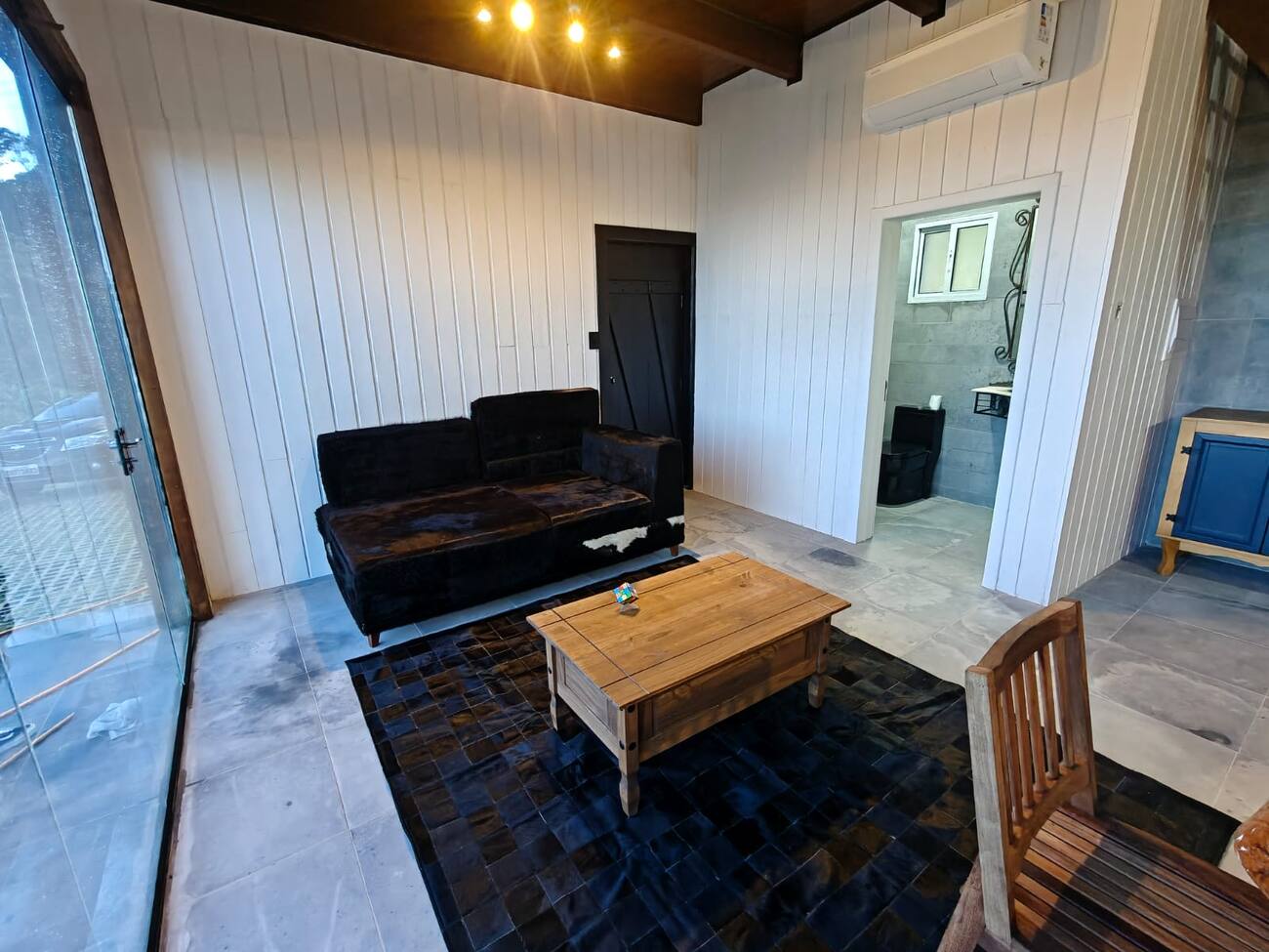 Sala de estar com sofá de couro preto, tapete de retalhos de couro e mesa de centro em madeira maciça, com porta de vidro para o jardim.