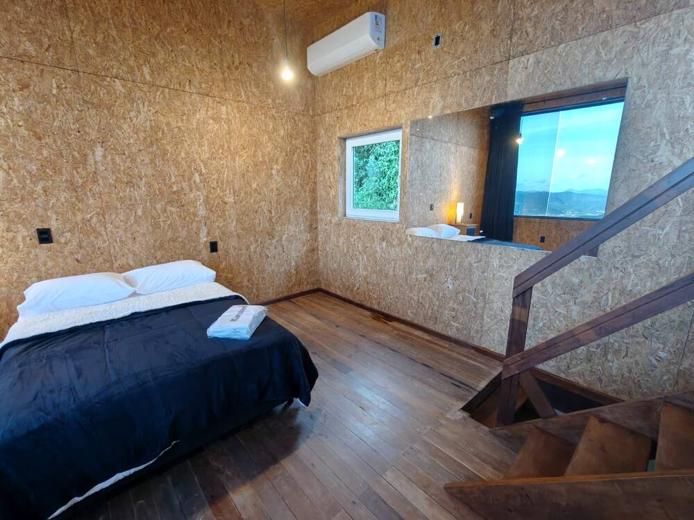 Cama de casal da suíte junto à janela, com espelho refletindo o quarto e escada de madeira descendo para o térreo.
