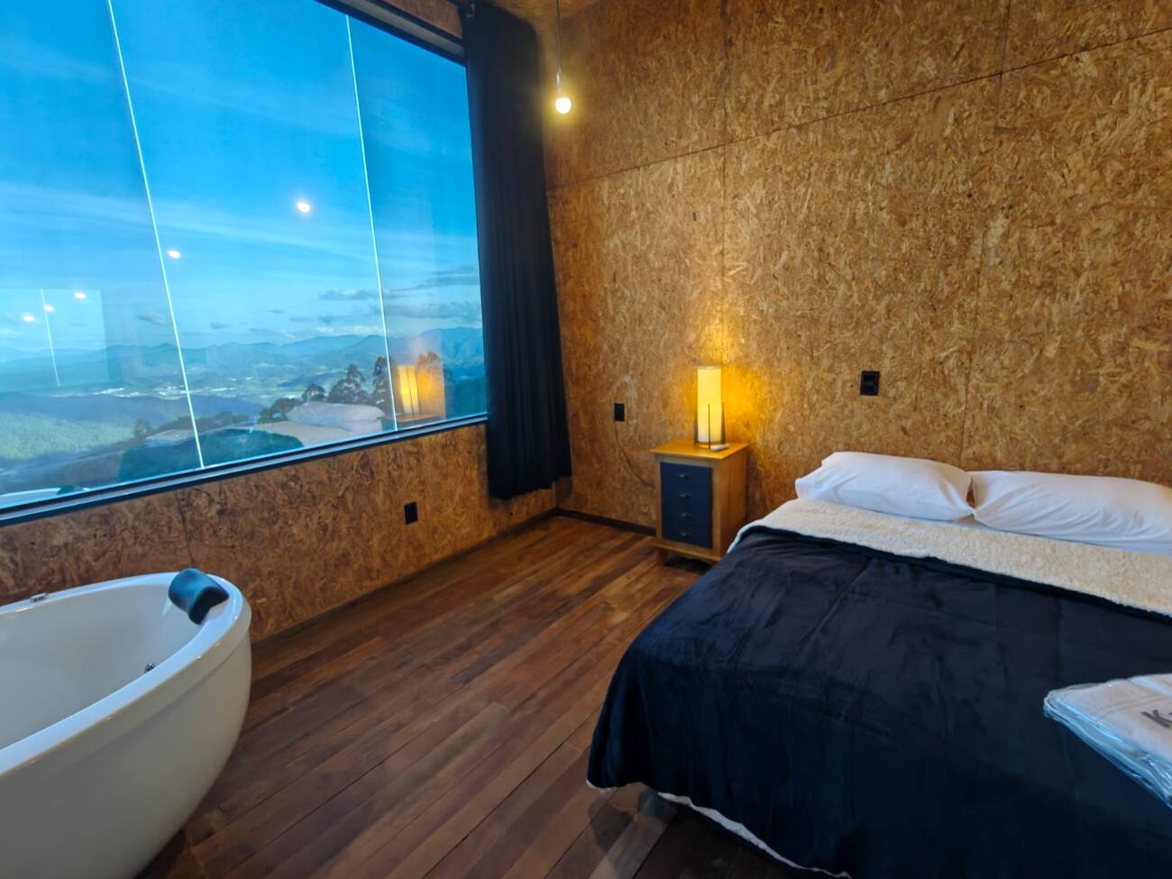 Quarto da suíte com cama de casal e banheira de hidromassagem, junto à janela panorâmica com vista para a serra ao entardecer.