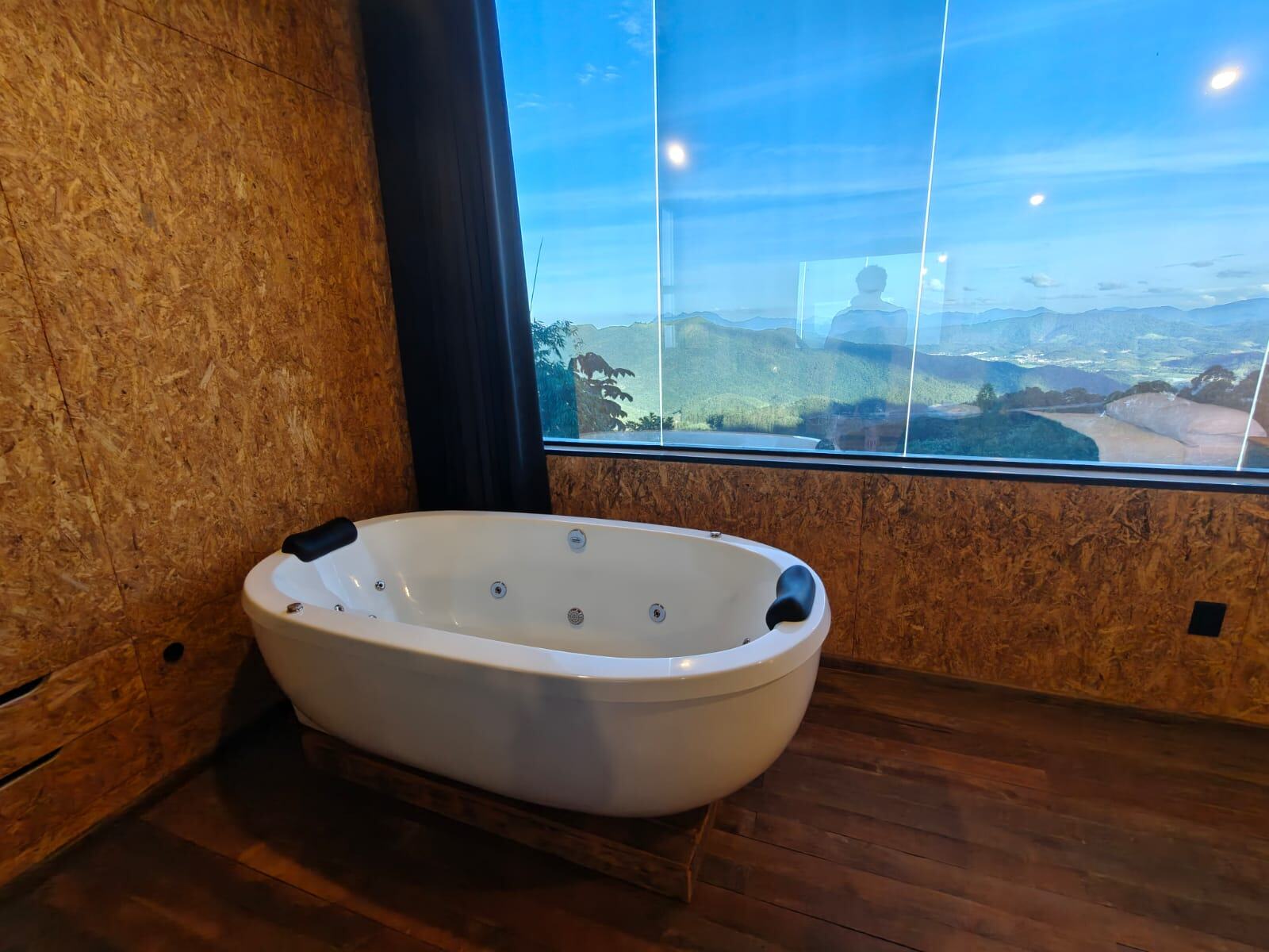 Banheira de hidromassagem em frente a uma janela do piso ao teto, com vista para as montanhas da Mata Atlântica ao entardecer.